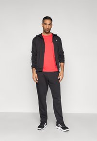 Čierna atletická bunda na zips s bielymi pruhmi, červené tričko, čierne tepláky a čierne tenisky s bielymi detailmi. Jednoduchý, priliehavý dizajn.