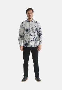 Chemise blanche avec motif abstrait navy, col boutonné, manches longues. Portée avec un pantalon sombre et des chaussures noires. Accent sur le design du motif unique.