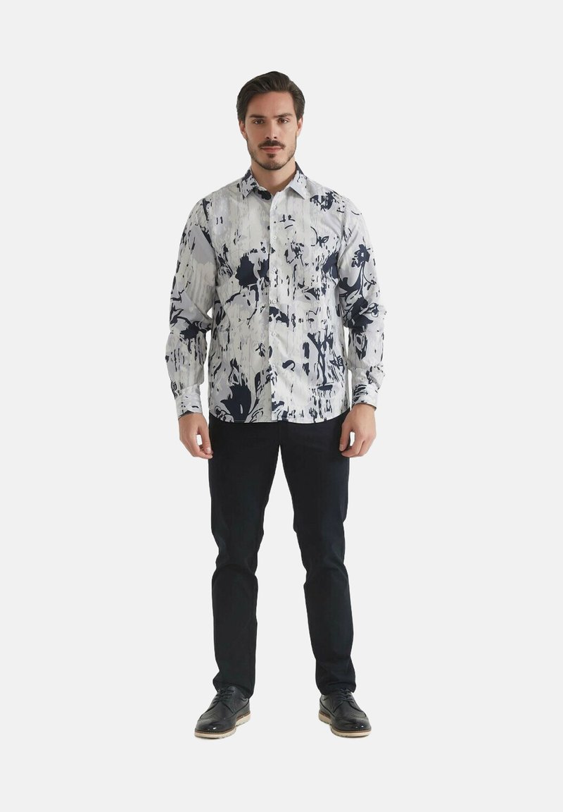 Chemise blanche avec motif abstrait navy, col boutonné, manches longues. Portée avec un pantalon sombre et des chaussures noires. Accent sur le design du motif unique.