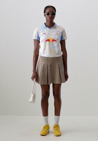 Polo shirt Puma avec un design abstrait coloré, col bleu ; jupe plissée beige ; lunettes de soleil blanches ; baskets jaunes ; chaussettes blanches ; petit sac blanc.