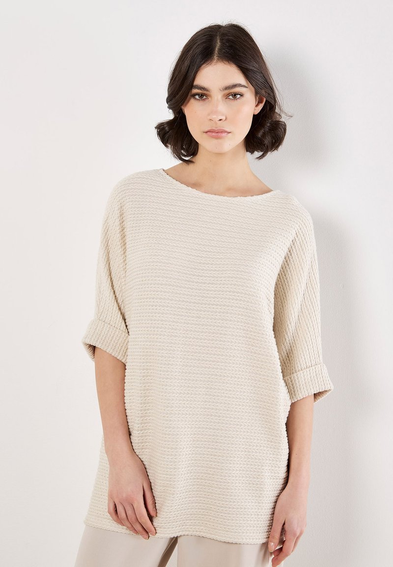 Světle béžový oversized pletený top s krátkými ohrnutými rukávy a texturovaným vzorem. Měkká a prodyšná tkanina, lodičkový výstřih, volný střih.