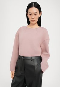 Lichtroze oversized trui met een ronde halslijn en subtiele naaddetails, gecombineerd met hooggewaiste zwarte leren broek.