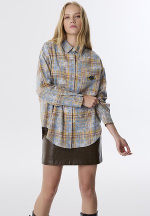 Camicia a quadri blu e gialla con maniche lunghe, chiusura con bottoni e tasca sul petto con logo. Indossata sopra una mini gonna in pelle marrone.