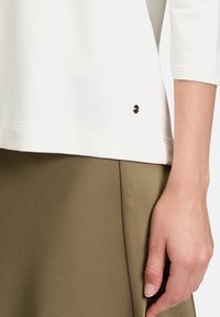 Nahaufnahme einer Hand neben olivgrüner Hose und einem weißen Langarmshirt mit einem kleinen schwarzen Knopfdetail.