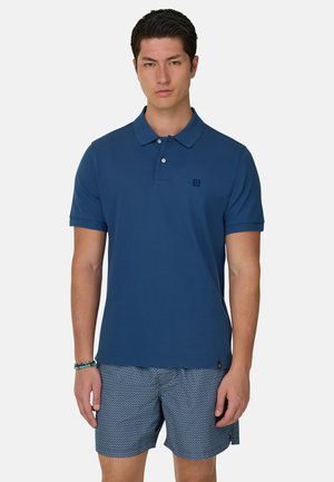 Hombre con una camisa polo de manga corta azul y pantalones cortos azules con estampado, de pie contra un fondo liso.