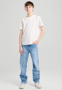 Weißes Baumwoll-T-Shirt kombiniert mit hellblauen Jeans. Das Model steht mit einer Hand in der Tasche und trägt helle Sneakers. Einfaches, lässiges Outfit.