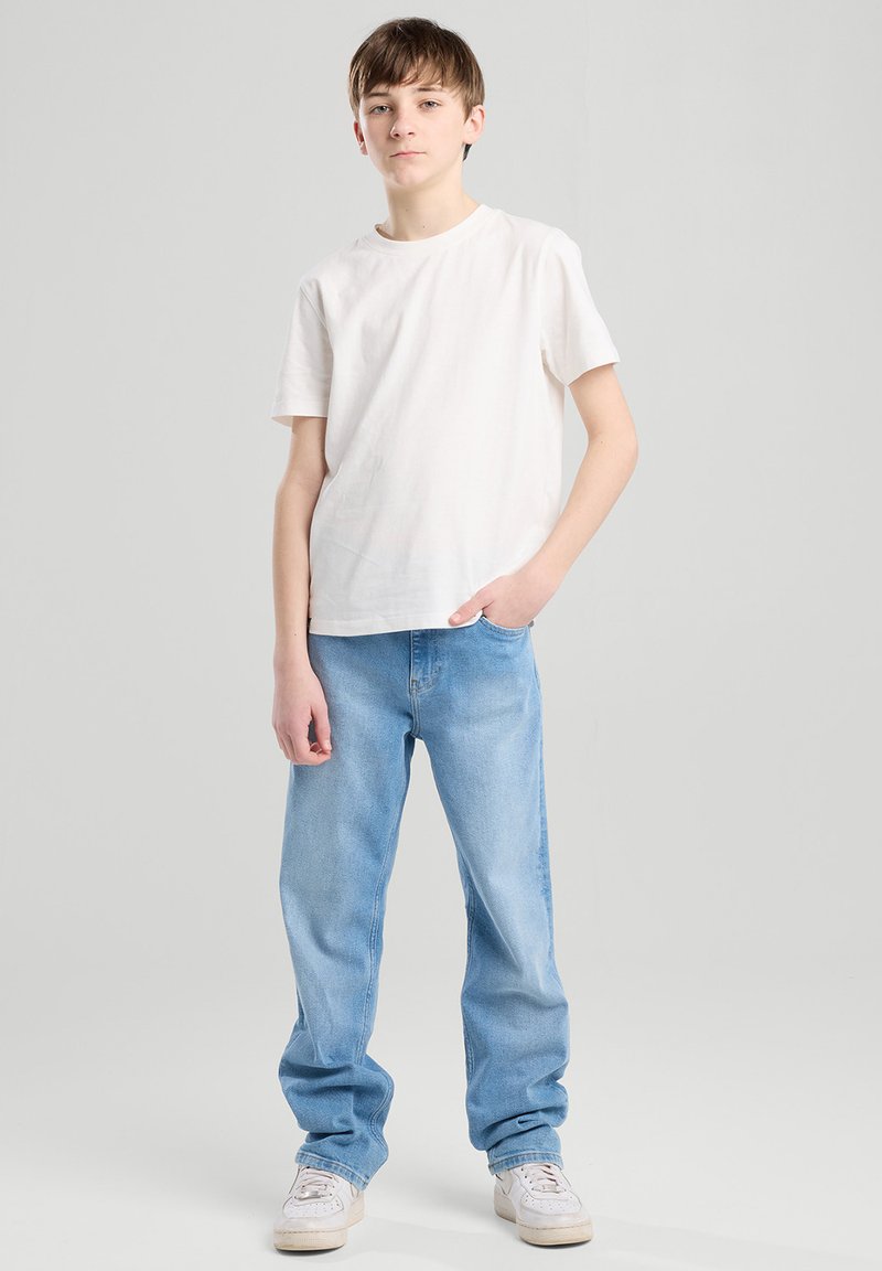 Weißes Baumwoll-T-Shirt kombiniert mit hellblauen Jeans. Das Model steht mit einer Hand in der Tasche und trägt helle Sneakers. Einfaches, lässiges Outfit.
