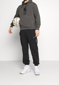 Grauer Hoodie mit NBA-Logo, dunkle Hose mit Seitentasche und weiße Sneakers. Das Modell hält einen Basketball und steht vor einem einfarbigen Hintergrund.