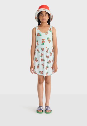 Meisje draagt een lichtblauw hemdje en een rok met oranje fruitprints, een rood-witte hoed, en blauwe sandalen met groene zolen.