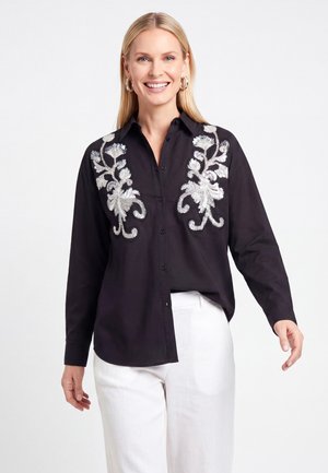 Femme souriante portant une chemise noire boutonnée avec une broderie florale argentée associée à un pantalon blanc sur un fond uni.
