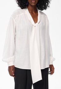 Kobieta ubrana w luźną białą bluzkę z długimi rękawami oraz wiązaną chustę przy kołnierzu, w komplecie z czarnymi spodniami, stoi na białym tle.