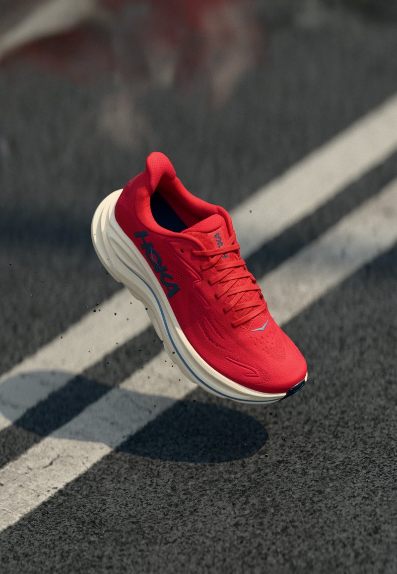 Κόκκινο παπούτσι τρεξίματος Hoka στον αέρα πάνω από δρόμο με άσφαλτο που έχει άσπρες διαγραμμίσεις και μ ικρά χαλικάκια.