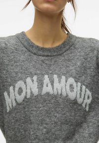 Pull gris en tricot avec un col rond, arborant le texte "MON AMOUR" en lettres blanches et texturées. Détail du tissu doux et chaud visible.