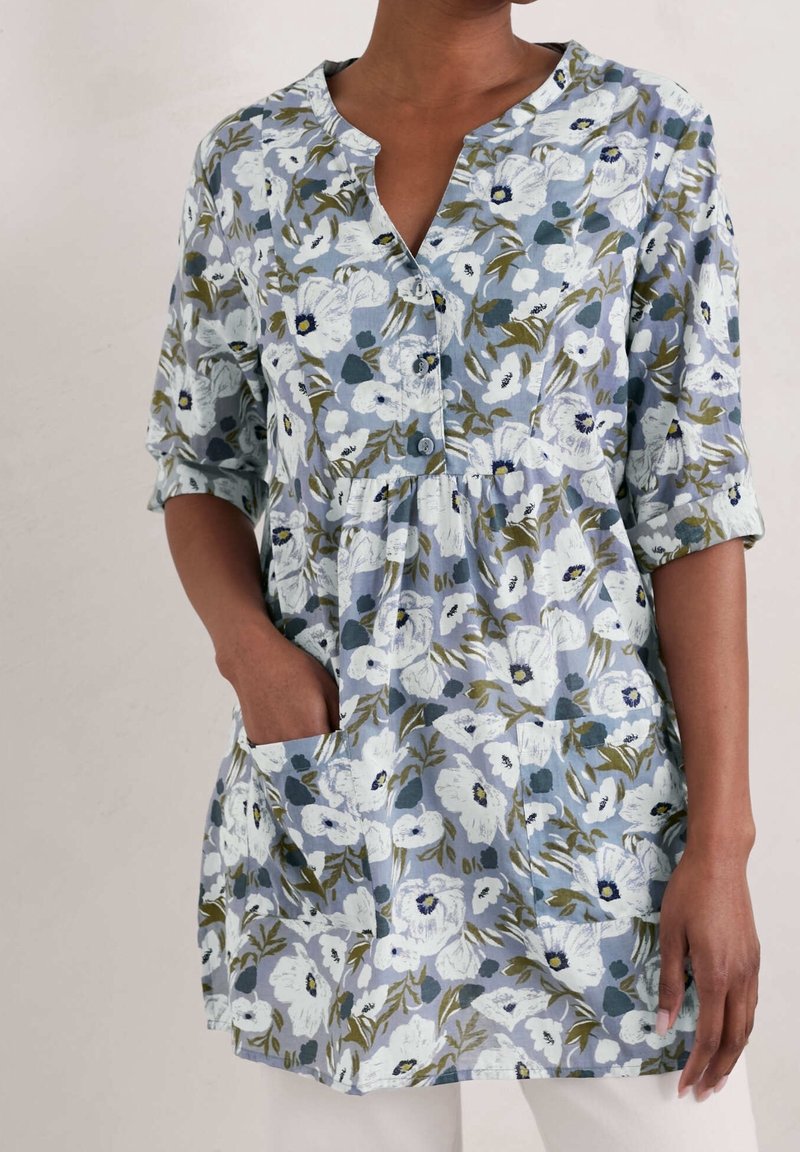 Femme portant une tunique bleue à fleurs boutonnée, manches retroussées et deux poches avant, main reposant dans une poche, sur un fond uni.