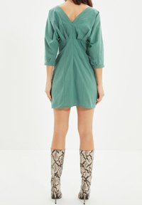 Vestido corto verde con escote en V en la espalda, mangas tres cuartos y detalle de cintura fruncida. Combinado con botas altas de serpiente hasta la rodilla.