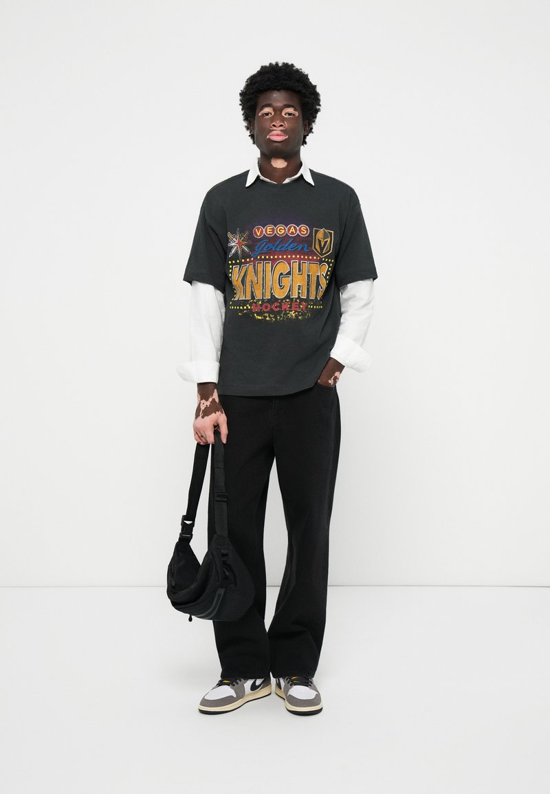 Junge Person mit Vitiligo, die ein schichtweises weißes Hemd und ein schwarzes T-Shirt der Vegas Golden Knights trägt, dazu schwarze Hose, Sneaker und eine schwarze Umhängetasche hält.