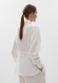 Chemise en lin blanc avec une taille ajustée et des manches décontractées, dotée d'un détail de couture au dos et de poches latérales. Texture lisse et respirante.