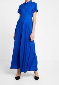 Sininen, lyhythihainen, napillinen mekko, jossa on tiukka yläosa ja liehuva hame. Valmistettu sileästä kankaasta, jossa on kaulus ja mustat kengät.