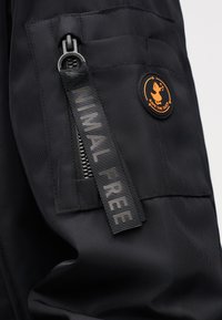 Manche de veste noire avec une poche zippée dotée d'une tirette "ANIMAL FREE" et un patch logo rond orange "SAVE THE DUCK".