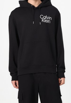 Sudadera con capucha negra de Calvin Klein con logo blanco en el pecho izquierdo, combinada con pantalones cargo negros, que cuentan con bolsillo frontal tipo canguro y capucha con cordón ajustable.