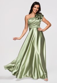 Femme posant dans une robe fluide en satin une épaule avec un grand détail floral vert clair, souriante sur un fond uni.