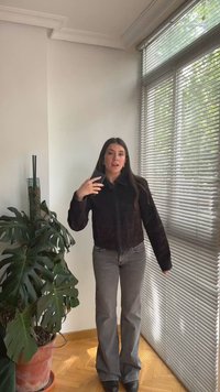 Νεαρή γυναίκα στέκεται σε εσωτερικό χώρο κοντά σε μεγάλο παράθυρο με περσίδες, κρατώντας ένα τηλέφωνο στο δεξί της χέρι, δίπλα σε ένα μεγάλο φυτώριο σε γλάστρα.