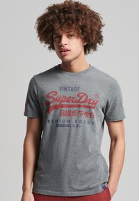 Jeune homme aux cheveux bouclés portant un T-shirt gris avec le logo vintage de SuperDry et un pantalon rouge, se tenant devant un fond clair.