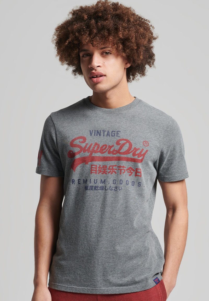 Jeune homme aux cheveux bouclés portant un T-shirt gris avec le logo vintage de SuperDry et un pantalon rouge, se tenant devant un fond clair.