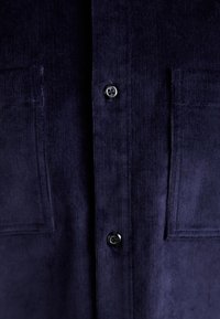 Camisa de pana marina con dos bolsillos frontales, cierre de botones y textura acanalada. Los botones son oscuros, redondos y tienen un ligero brillo.