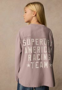 Ilga rankovė levandų spalvos marškiniai su "SUPERDRY AMERICAN RACING TEAM" dideliais baltais įtrūkusiais rašmenimis atvirkščiai, šonuose įsiūtos skeltukai.