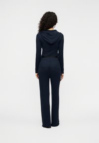 Φόρμα με κουκούλα σε ναυτικό μπλε χρώμα, μακριά μανίκια, εφαρμοστή μέση και παντελόνι με φαρδιά μπατζάκια· λεία υφή με διακριτικές ραφές.