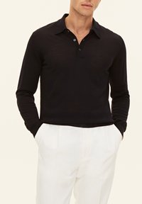 Långärmad svart pikétröja med krage, tvåknapplöstning och en figurnära design. Bärs med vita byxor som visar en strukturerad tyg.