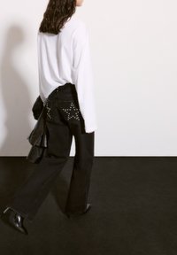 Svarta denimjeans med nitar på fickorna, kombinerat med en vit oversized långärmad skjorta och svarta klackstövlar. Långt hår synligt.