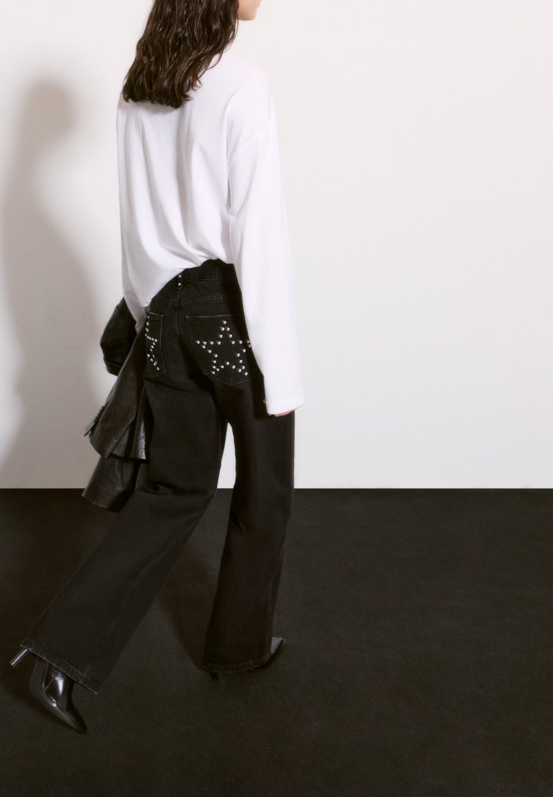 Svarta denimjeans med nitar på fickorna, kombinerat med en vit oversized långärmad skjorta och svarta klackstövlar. Långt hår synligt.