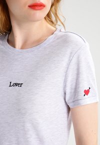 Camiseta de algodón gris con el texto negro bordado "Lover" en el frente y un corazón rojo con flechas en la manga. Mangas cortas, corte informal.