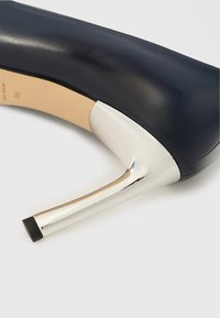 Marineblå skinnhøyhæl med en smal, sølvfarget metallic hæl og sort gummitupp. Jevn tekstur som fremhever det elegante designet.