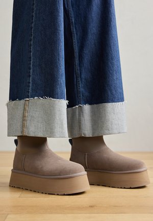 Botines de plataforma gruesa en ante topo combinados con vaqueros de pierna ancha y puños en denim azul sobre un suelo de madera.