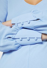 Světle modrá košile s dlouhými rukávy, které mají nabíranou látku a tři knoflíky potažené látkou na manžetách. Hladká textura, výstřih ve tvaru V.