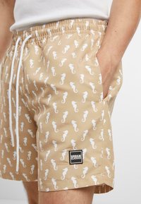 Beige színű úszóshort, fehér tengeri lovacskák mintájával, rugalmas derékpánttal, állítható zsinórral és a hemnél egy fekete címkével, amelyen az "URBAN CLASSICS" felirat olvasható.
