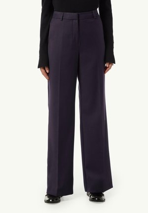 Weite Stoffhosen in tiefem Marineblau mit glatter Oberfläche, hoher Taille, Bundfalten und seitlichen Taschen. Glänzende schwarze Schuhe runden den Look ab.