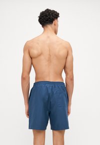 Marineblaue Schwimmshorts mit einem elastischen Taillenbund und einer Gesäßtasche, hergestellt aus einem glatten, leichten Material. Model von hinten gesehen.