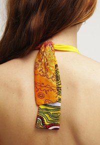 Des cheveux bruns tombent sur le dos d'une personne portant un haut halter jaune noué avec un foulard à motifs multicolores orange et jaune.