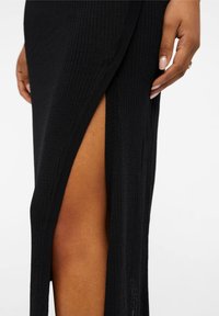 Robe noire en maille côtelée avec une fente latérale haute, une texture douce et une coupe ajustée. Peau visible à travers la fente.