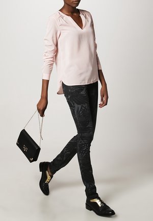 Helles rosa V-Ausschnitt-Bluse mit langen Ärmeln, kombiniert mit schwarzen, gemusterten Skinny-Jeans, schwarzen Schuhen mit goldenen Akzenten und einer schwarzen Handtasche.