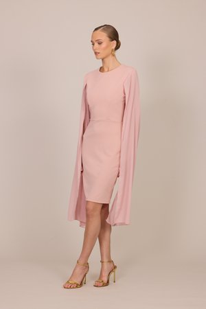 Femeie purtând o rochie roz-pal cu lungimea până la genunchi, mâneci lungi, ample, tip pelerină și sandale aurii cu toc înalt, stând în fața unui fundal simplu.