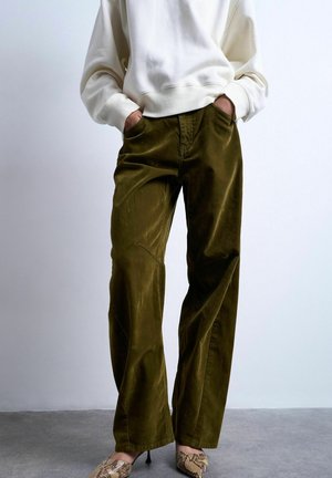 Person iført olivengrønne vidde bukser, hvid sweatshirt og beige højhælede sko med slangeprint, står med hænderne i lommerne.