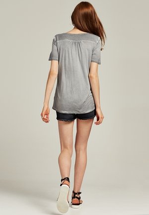 Gray short-sleeve top with a relaxed fit and gathered detail at the back, paired with dark denim shorts and flat sandals.