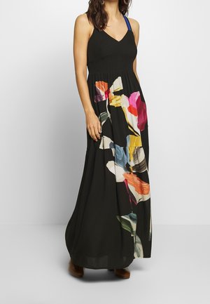 Femme portant une robe maxi noire sans manches avec des imprimés floraux abstraits colorés et un détail de bretelle bleue, debout devant un fond uni.