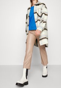 Camisa de cuadros crema de gran tamaño con rayas azules y negras, combinada con pantalones de cuero sintético beige y botas de tacón cuadrado blancas con detalles negros.