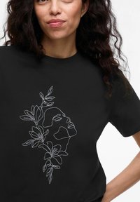 Svart t-shirt med vit konturbroderi av ett ansikte som är sammanflätat med blommiga element, vilket framhäver den intrikata designen och texturen.